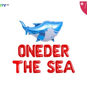 May include: A blue and white shark-shaped balloon floats above red balloon letters spelling "ONEDER THE SEA." The image includes a balloon straw and string, perfect for a first birthday party. The party supply is on a white background.