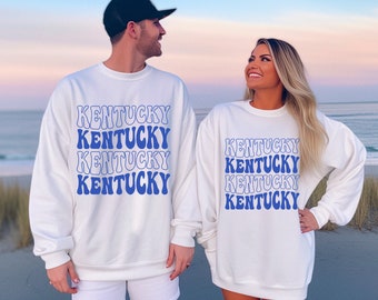 May include: Two white sweatshirts with the word "Kentucky" repeated in a blue retro font. The sweatshirts are worn by two people standing on a beach.