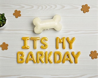 May include: A festive arrangement featuring a cream-colored bone-shaped balloon and gold foil letter balloons spelling "ITS MY BARKDAY." The scene includes brown paw-shaped cutouts and a small potted plant, all set on a light wood-grain surface.