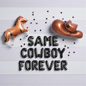 May include: A brown horse and a copper-colored cowboy hat balloon are displayed with black balloon letters that spell "SAME COWBOY FOREVER". Black star and heart confetti are scattered on a white wooden background.