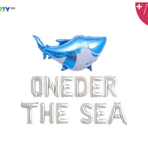 May include: A blue and white shark-shaped balloon floats above silver foil letter balloons spelling "ONEDER THE SEA." The image includes a balloon straw and string, perfect for a first birthday party with a nautical theme.