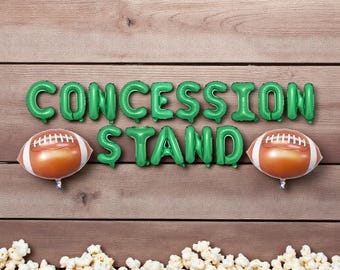 May include: Green foil balloon letters spelling "CONCESSION STAND" with two football-shaped balloons. The balloons are arranged on a wooden background with a popcorn border at the bottom. A festive theme.