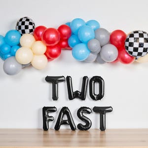 May include: A festive balloon garland in shades of blue, red, cream, and gray, with two checkered flag balloons. Below, black foil balloons spell out "TWO FAST", perfect for a race car themed birthday party.