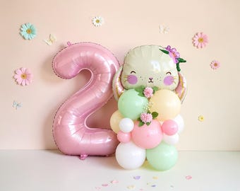 May include: A large pink foil balloon in the shape of the number two next to a balloon arrangement featuring a light green bunny face balloon with floral accents, surrounded by smaller round balloons in shades of white, pink, and light green.