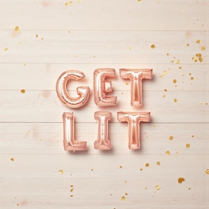 以下が含まれることがあります： 明るいピンク色の木製の背景に、「GET LIT」と書かれたローズゴールドのホイルバルーンと、金色の紙吹雪が散りばめられています。 バルーンは膨らませて、単語を形成するように配置されています。