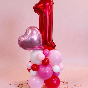 May include: A festive balloon arrangement for a first birthday celebration. It features a large, shiny red number one balloon, a pink heart-shaped balloon, and a cluster of pink, red, and white balloons. Confetti is scattered around the base.