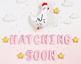 May include: A white chicken-shaped foil balloon with a red comb and pink cheeks, surrounded by small white cloud balloons, gold star balloons, and pink letter balloons spelling out the phrase HATCHING SOON against a light background.