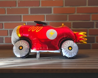 May include: A red and gold foil balloon shaped like a race car. The car has silver wheels with yellow accents, a black steering wheel, and a white circle with a yellow lightning bolt. The car has the number "000" in yellow.