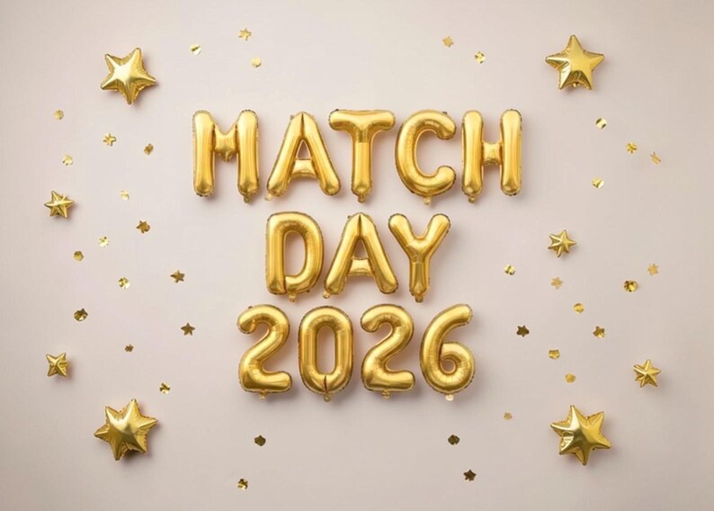 May include: Gold foil balloons spell out "MATCH DAY 2026" on a light beige background, with gold star-shaped confetti and star balloons scattered around. The balloons are inflated and shiny.