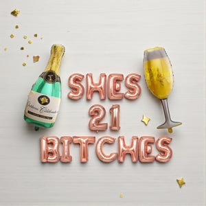 May include: A festive arrangement of rose gold foil balloons spelling "SHES 21 BITCHES" with a champagne bottle and glass balloon. The champagne bottle is green with gold accents, and the glass is filled with yellow liquid.