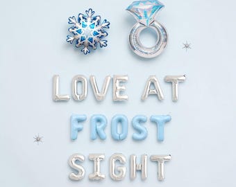 May include: Silver and light blue foil balloons spelling out LOVE AT FROST SIGHT, accompanied by a blue snowflake balloon and a silver diamond ring balloon. The letters are arranged in three rows against a light blue background.