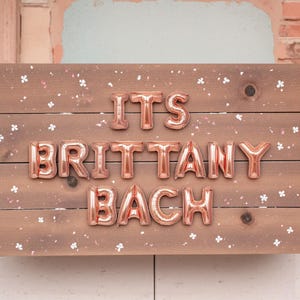 May include: A wooden sign with the phrase "ITS BRITTANY BACH" spelled out in rose gold balloon letters. The sign is decorated with small white flower accents and a light pink background.