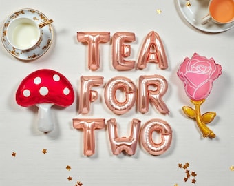 以下が含まれることがあります： 「TEA FOR TWO」と書かれたローズゴールドの風船を使ったティーパーティーの華やかなアレンジ。赤いキノコの風船、ピンクのバラの風船、2つのティーカップ、ソーサー付きのティーカップが含まれています。金色の星の紙吹雪が散らばっています。