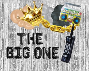 May include: A party backdrop with silver fringe, black balloon letters spelling "THE BIG ONE", and a gold crown balloon. Other balloons are tan, gray, and black. A gold chain connects the crown to a boombox and a black cell phone balloon.