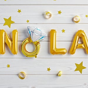 May include: Gold foil balloons spelling "NOLA" with a diamond ring balloon in the center, set against a white wooden background. Gold star confetti and ribbon accents complete the festive arrangement.