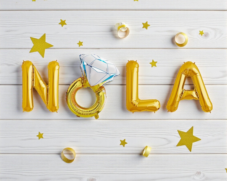 May include: Gold foil balloons spelling "NOLA" with a diamond ring balloon in the center, set against a white wooden background. Gold star confetti and ribbon accents complete the festive arrangement.