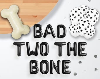 May include: A collection of party decorations, including a bone-shaped balloon, black letter balloons spelling "BAD TWO THE BONE", and white balloons with black paw prints. Silver confetti dots the background.