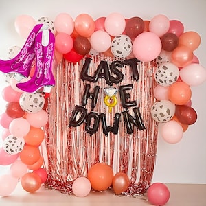 May include: A festive party decoration set with pink, peach, and brown balloons, a metallic fringe backdrop, and foil balloon letters spelling "LAST HOE DOWN". Includes a pair of pink cowboy boot balloons and a ring balloon.