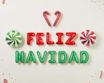 May include: A set of foil balloons spelling FELIZ NAVIDAD in red and green, accompanied by two round peppermint candy balloons and two candy cane balloons, with small decorative pine branches and confetti scattered around.