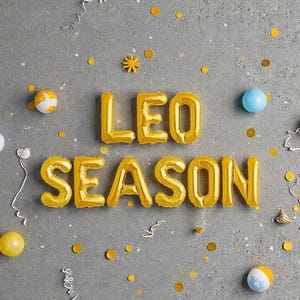 May include: Gold foil balloon letters spell "LEO SEASON" on a gray surface, surrounded by confetti, small decorative balls, and streamers. The balloons are inflated and arranged to create a festive display.