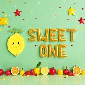May include: A festive scene with a green background, featuring a yellow lemon balloon with a smiling face, gold foil balloons spelling "SWEET ONE", and various fruits. Red and yellow star-shaped decorations and scattered confetti add to the celebratory atmosphere.