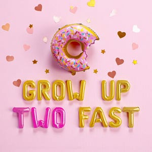 May include: A pink background featuring a donut-shaped balloon and gold letter balloons spelling "GROW UP TWO FAST". Heart and star-shaped confetti is scattered around. The donut balloon is pink with colourful sprinkles.