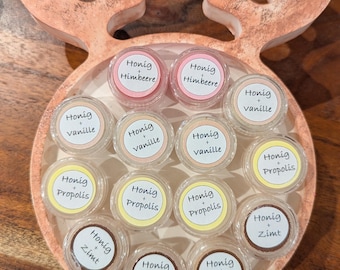 Könnte beinhalten: Ein Holztablett in Form eines Rentiers mit 12 kleinen Honiggläsern. Die Gläser sind mit verschiedenen Geschmacksrichtungen beschriftet, darunter Vanille, Himbeere, Zimt und Propolis.