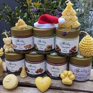 Könnte beinhalten: Sortiment an Honig-Duftkerzen in Glasbehältern mit verschiedenen weihnachtlichen Dekorationen, darunter ein Rentier, eine Tannenzapfen, ein Weihnachtsmann und ein Weihnachtsbaum. Die Behälter sind mit dem Honiggeschmack beschriftet, wie "Nougat in Honig" und "Marzipan in Honig".