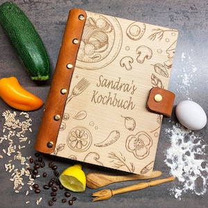 Pode incluir: Um livro de receitas encadernado em couro castanho com uma capa de madeira. A capa é gravada com um desenho de vários alimentos, incluindo massa, pimentões, limões e flores. O texto "Sandra's Kochbuch" está gravado na capa.