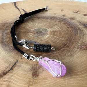 May include: A handmade necklace with a bright pink crystal encased in silver wire. It features a black cord, black lava beads, and a silver feather charm. The jewellery is displayed on a wooden surface.
