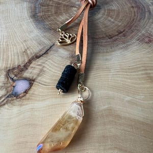 May include: A handmade pendant featuring a citrine crystal, black bead, and gold lotus charm. The pendant hangs from a brown leather cord with a knotted top. The citrine crystal is a light orange colour.