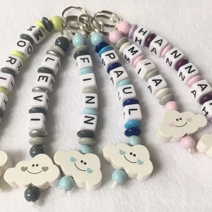 Könnte beinhalten: Fünf bunte Perlen-Schlüsselanhänger mit verschiedenen Namen, die darauf geschrieben sind. Jeder Schlüsselanhänger hat einen kleinen, hölzernen Wolken-Anhänger mit einem Smiley und zwei Herzen.