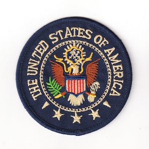 May include: A blue embroidered patch with the words "The United States of America" in gold lettering around a gold eagle with a shield and olive branch.