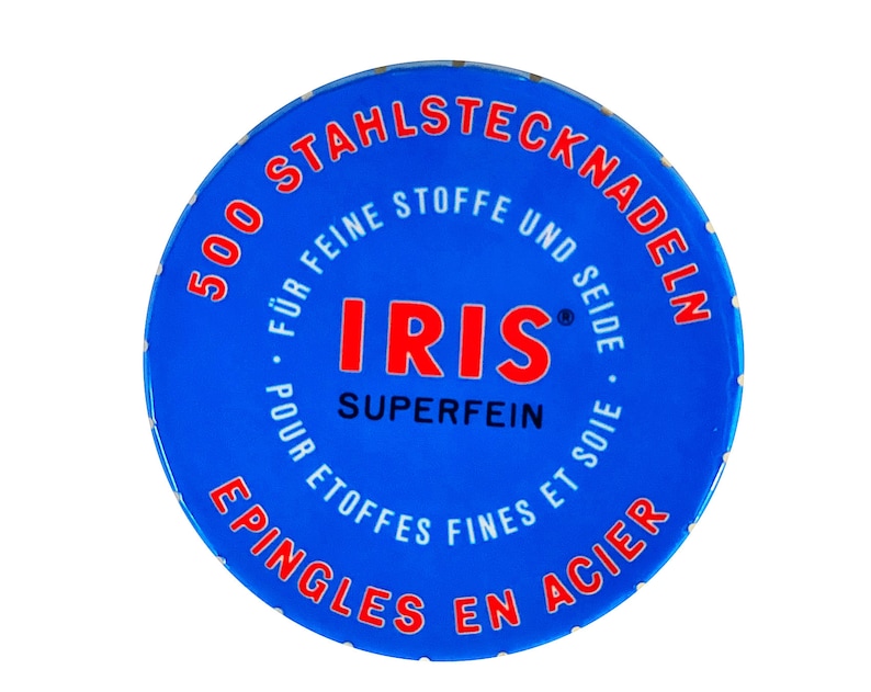 Peut inclure: Une bo&icirc;te ronde bleue avec des inscriptions blanches qui disent "500 Stahlstecknadeln" et "Iris Superfein" en rouge. La bo&icirc;te est pour des aiguilles &agrave; coudre.