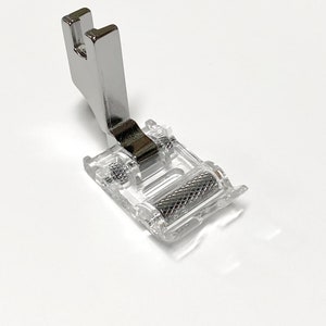 May include: A clear, plastic sewing machine foot with a metal shank and a textured roller. The foot is designed to help guide fabric smoothly under the needle. The metal shank is silver and the roller is clear.