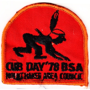 May include: A red embroidered patch with an orange border. The patch features a black silhouette of a person in a headdress, kneeling with a feather in their hand. The text "Cub Day '78 BSA Mohawk Area Council" is embroidered in black below the figure.