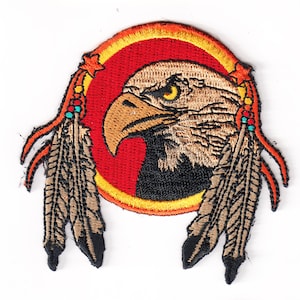May include: An embroidered patch featuring a brown and black eagle head with a yellow eye, surrounded by a red circle with a yellow border. Two brown feathers with orange and blue accents are on either side of the eagle.