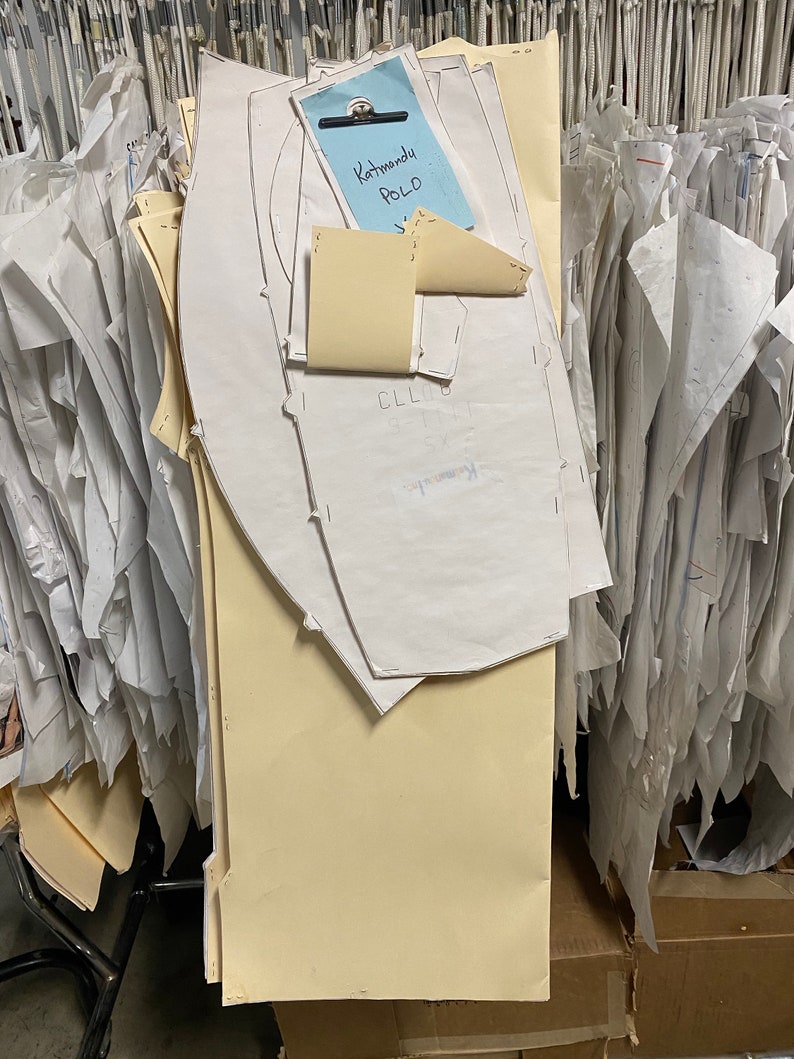 Op de afbeelding: Een stapel witte en cr&egrave;mekleurige papieren patronen voor kleding, met een blauwe notitie bovenop die "Kathmandu POLO" zegt.