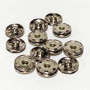 May include: A close-up of a group of silver metal snap fasteners. The fasteners are arranged in a circular pattern on a white surface.
