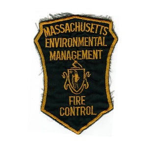 May include: A dark green patch with gold embroidered text that reads "Massachusetts Environmental Management Fire Control". A gold embroidered crest with a star and a hand holding a torch is in the center of the patch.