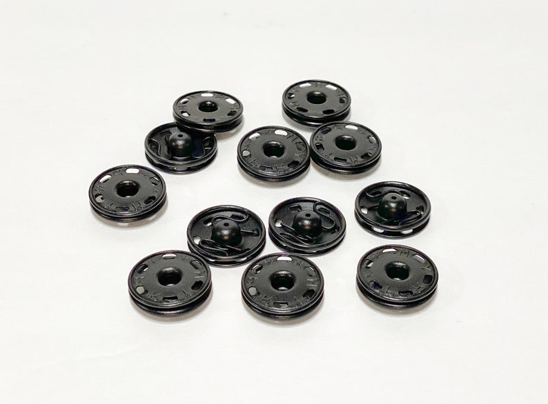 K&ouml;nnte beinhalten: Nahaufnahme von 10 schwarzen Kunststoff-Druckkn&ouml;pfen. Die Kn&ouml;pfe sind rund und haben eine erhabene Mitte mit einem kleinen Loch in der Mitte. Die Kn&ouml;pfe sind in einem zuf&auml;lligen Muster auf einer wei&szlig;en Oberfl&auml;che angeordnet.
