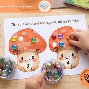 Könnte beinhalten: Ein weißes Lernblatt mit zwei aquarellartigen Pilzen, auf deren Stielen die Zahlen 3 und 4 stehen. Der Text lautet Zähle die Glassteine und lege sie auf die Punkte. Bunte Glassteine sind auf den Pilzhüten platziert, um die Mengen zu veranschaulichen.