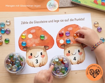 May include: A white educational worksheet featuring two watercolor-style mushrooms with the numbers 3 and 4 printed on their stems. The text reads Zähle die Glassteine und lege sie auf die Punkte. Colorful glass pebbles are placed on the mushroom caps.