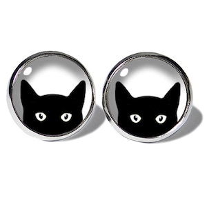 Könnte beinhalten: Ein Paar silberne Ohrstecker mit einem schwarzen Katzenkopf-Design. Die Augen der Katze sind weiß und die Katze schaut aus einem weißen Kreis heraus.