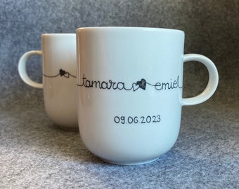 Könnte beinhalten: Zwei weiße Keramikbecher mit schwarzem handgeschriebenem Text. Ein Becher hat die Namen "tamara emiel" und das Datum "09.06.2023" mit Herzmotiven. Der andere Becher hat ein Herzmotiv. Beide Becher haben einen gebogenen Griff.