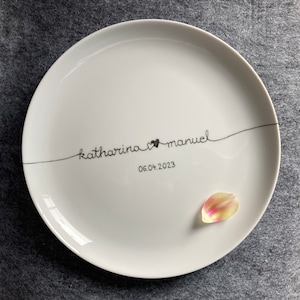 Könnte beinhalten: Weiße Keramikplatte mit schwarzem handgeschriebenen Text "katharina & manuel" und dem Datum "06.04.2023". Ein rosa Blütenblatt liegt auf dem Teller.