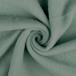 Può includere: Primo piano di un tessuto morbido di colore verde salvia con una superficie testurizzata e pelosa. Il tessuto è piegato e drappeggiato, creando un motivo a spirale.