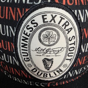 May include: Black baseball cap with a repeating "Guinness" pattern in red and white. The cap features a circular patch with "Guinness Extra Stout" text, a shamrock, and a harp symbol.
