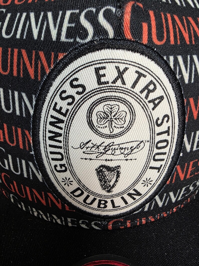 May include: Black baseball cap with a repeating "Guinness" pattern in red and white. The cap features a circular patch with "Guinness Extra Stout" text, a shamrock, and a harp symbol.