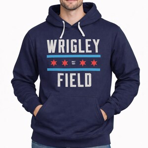 May include: Navy blue hoodie with the text "WRIGLEY FIELD" in white block letters. Above the text are two horizontal blue lines and three red stars. The hoodie has a front pocket and a white drawstring.